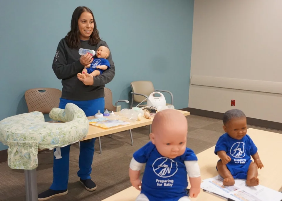Woman holding infant doll while speaking to class.