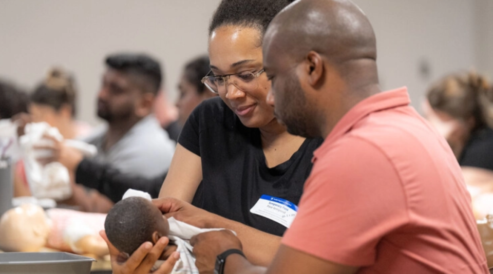 Two people holding an infant doll while engaging in a hands-on workshop to teach skills.